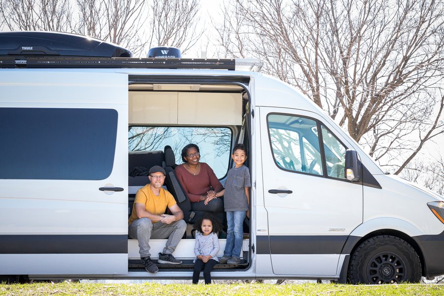 Family of four sitting by a camper van on a sunny day, with trees in the background.